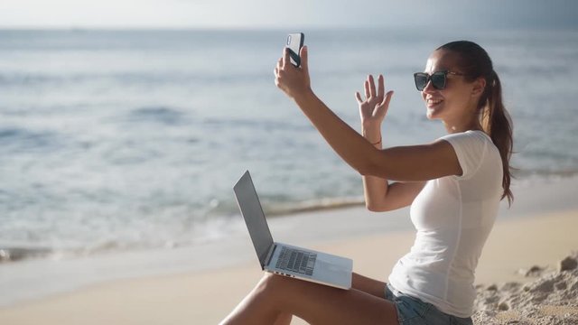 Woman with laptop sits on beach near ocean and uses phone for video call