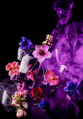 A glass perfume bottle shatters and bright spring flowers and clouds of blue and purple vapor burst out of it against a dark background