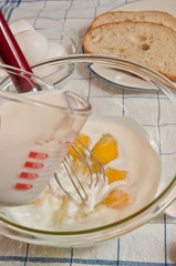 Top view, close distance of a glass bowel of raw, local, eggs and yolks, with a metal whisk, red wood handle,  glass measuring glass, slices of bread on a blue, white towel