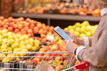 Mature female customer scrolling through list of products to buy in supermarket