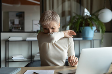 Front view young female manager with short haircut coughing or sneezing in elbow while working on...