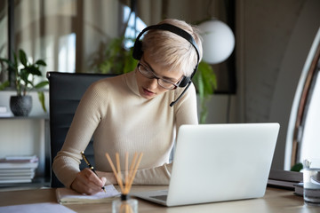 Focused business woman in glasses wearing headset with microphone sitting at desk, listening to...