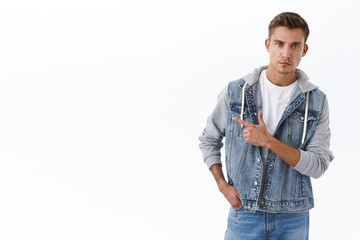 Serious-looking confident blond man in denim jacket, pointing finger left and looking focused with determined face camera, recommend try item, subscribe or click link of banner on white blank space