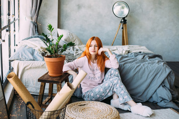 A redheaded pretty girl in pink pajamas is sitting next to the bed - 335599952