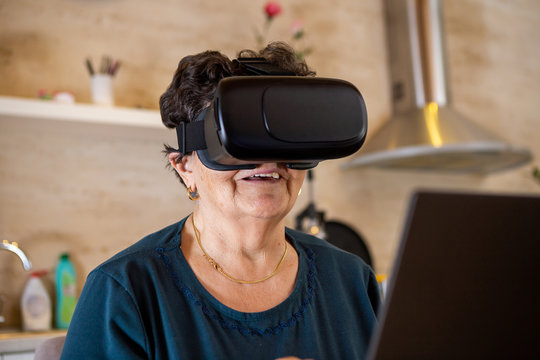A senior woman with black short hair in a turquoise T-shirt sits in front of a laptop and has virtual reality glasses on her head. She is at the table in the kitchen.