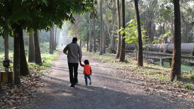 Backside of an Indian brunette father and his baby boy in winter garments walking in a forest path in winter afternoon in natural green background. Indian lifestyle and parenthood.
