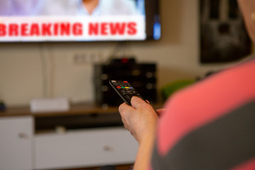 A senior woman sits in front of the television watching television. She holds a remote control in her hand that she points at the TV because she wants to change the channel. 