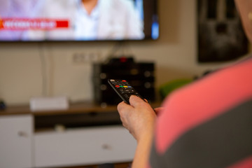 A senior woman sits in front of the television watching television. She holds a remote control in her hand that she points at the TV because she wants to change the channel. 