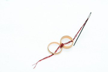 On a white background two gold wedding rings are connected by a red thread coming from a sewing needle. Concept - betrothal, wedding, love for life