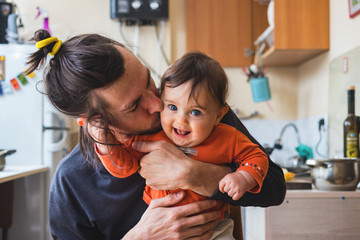 Hipster young father hug and kiss his baby infant boy holding on hands. At home on kitchen...
