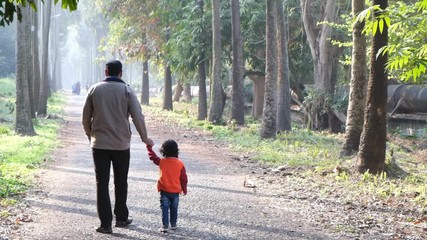 Backside of an Indian brunette father and his baby boy in winter garments walking in a forest path in winter afternoon in natural green background. Indian lifestyle and parenthood.