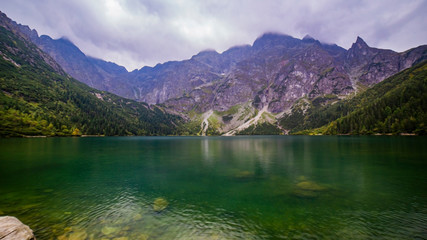 Naklejka na ściany i meble Scenic view of foggy mountains cover by dark clouds and green forest with a reflection in a lake. Stony shore. Morskie Oko. Marine Eye. High Tatras, Zakopane, Poland Concept of nature and tourism