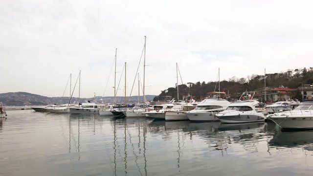 Cloudy foggy day. Private yachts and sailing boats with lowered sails standing on pier in bay. Reflections in water. People walk along promenade. Turkey, Istanbul, Tarabya, March 2020.