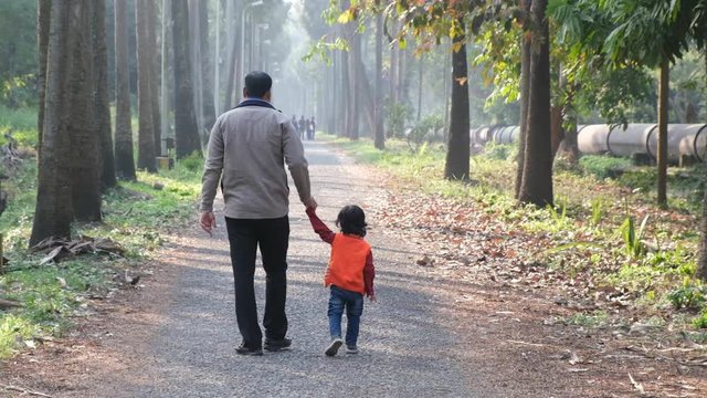 Backside of an Indian brunette father and his baby boy in winter garments walking in a forest path in winter afternoon in natural green background. Indian lifestyle and parenthood.