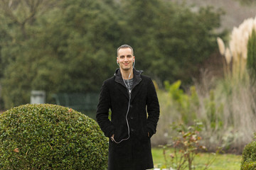 Young Man Wearing Coat and Listening to Earphones Posing in Garden