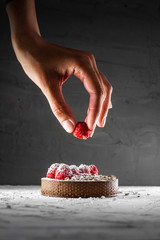 Appetizing tart with dark chocolate, crushed nuts, fresh raspberries and powdered sugar, chef's hand, decorates it, putting a berry, dark concrete textured background, natural color grade, copy space.