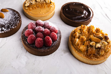 Delicious tarts with caramel and hazelnuts, with dark chocolate, raspberries, almond and powdered sugar on the light colored background. Natural light and color grade. Copy space