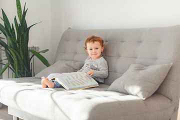 A little girl about 2 years old. In a light interior of neutral colors, reads a big book, backlight.