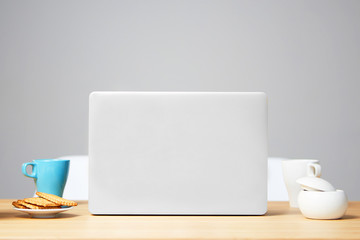 Image of open laptop on wooden table in front of grey background. Blue cup and cookies on the table. For mockup