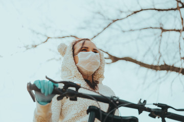 Portrait of young woman in medical mask and gloves with bicycle against sky. Adult female covering face to protect yourself from diseases on walk. Concept of threat of coronavirus epidemic infection.