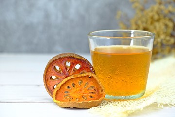 Cold bael fruit juice herbal drink served in a glass surrounded by dried pieces of bael fruit on white wooden table with concrete background.