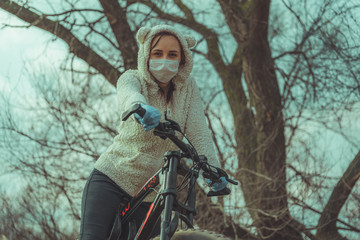 Fototapeta na wymiar Young woman in medical mask and gloves stands with bicycle, holding on to rudder in countryside. Female protecting yourself from diseases on walk. Concept of threat of coronavirus epidemic infection.
