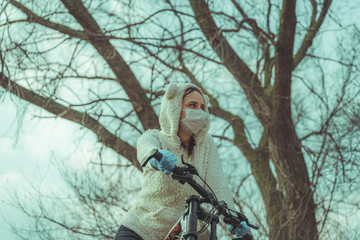 Young woman in medical mask and gloves stands with bicycle, holding on to rudder in countryside. Female protecting yourself from diseases on walk. Concept of threat of coronavirus epidemic infection.