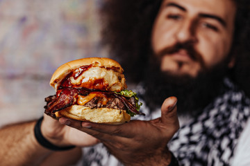 Curly hipster holding  craft mouth-watering bacon and angus beef burger flavored with crunchy fried onion, adding freshly cut potato fries on the side recommended for meat lovers. - 335618186