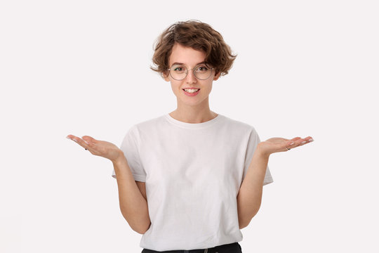 Clueless young woman in a white shirt and eyeglasses shrugging shoulders being at loss.