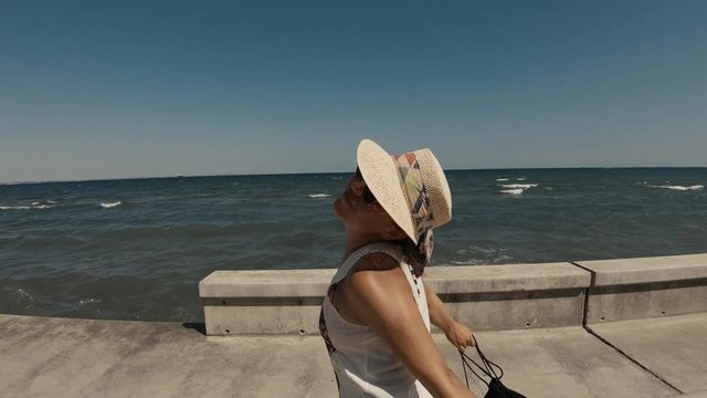 A young happy girl in her twenties wearing a hat and sunglasses, playing and turning around on a seaside dock. Enjoying summer in Cyprys with a wavy sea in the background on a sunny day.