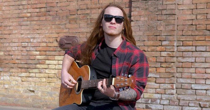 Young male with long hair and tattoo play on acoustic guitar outdoors on the street