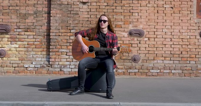 Young male with long hair and tattoo play on acoustic guitar outdoors on the street