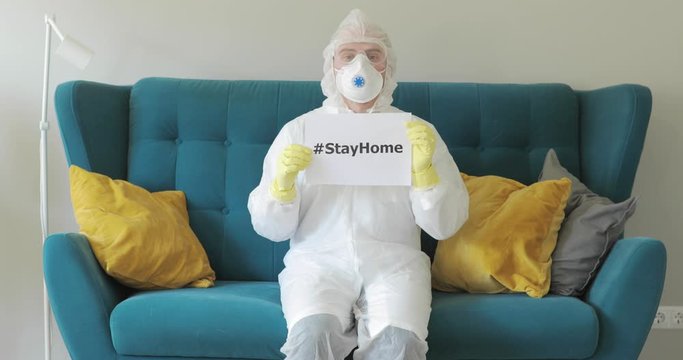 Doctor man in protective suit with inscription hashtag stay at home. He is sitting on sofa. Mask, respirator, glasses, suit during coronavirus pandemic, epidemic. Stayhome to fight with COVID-19 sars.