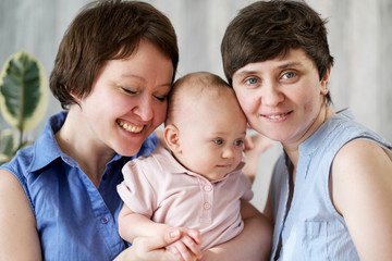 A young gay lesbian couple with a baby in their arms. They're posing at home.