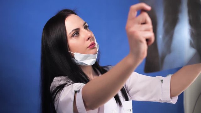 Female doctor examines x-ray of lungs, holding it in hands indoors. Experienced expert dressed in white medical.Blue background.