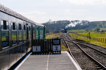 Steam train approaches a platform with another train stationary.