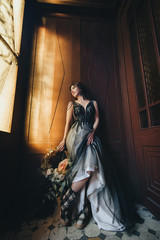 Pretty bride in a long dress with a wreath and flowers stands in the old interior with a wooden door and window. Wedding portrait of a beautiful girl. Photography, concept.