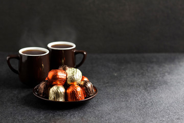 Chocolate candies wrapped in multicolored foil and two cups of coffee on black background