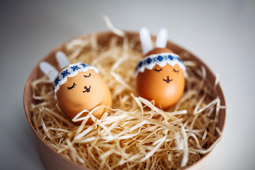 Two little easter eggs with a painted drawn rabbit faces and ears on them laying in hay on the table isolated. Easter holidays decorations and preparations concept. Holy religious day. Copyspace.