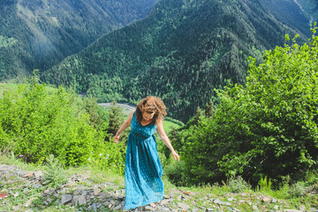 Elegant woman in blue dress at the mountains. Happy girl with wavy hair. Freedom concept. The background of the majestic mountains of the Caucasus and wooden fence. Svaneti, Georgia.