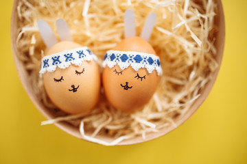 Two little easter eggs with a painted drawn rabbit faces and ears laying on the yellow table in hay isolated. Easter holidays decorations, preparations concept. Holy religious day. Copyspace.