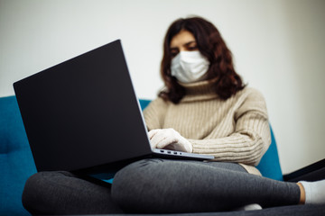 Fototapeta premium Young woman working from home during quarantine due to coronavirus pandemia. Beautiful girl stays home wearing medical mask and gloves and typing on a laptop. Covid-19 epidemia worldwide concept.