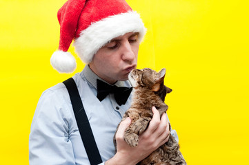 handsome man in red christmas santa hat, blue shirt and black suspender holding and kissing cute brown tabby cat in front of yellow background
