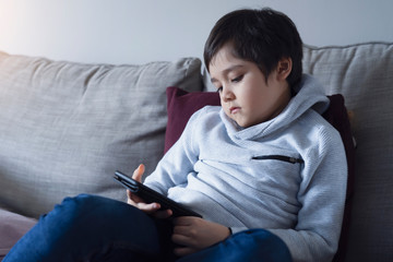 Kid self isolation using cellphone for homework, Sad child sitting on sofa looking down deep in thought,Boy learning on internet from home during school of. Home schooling, learning online education