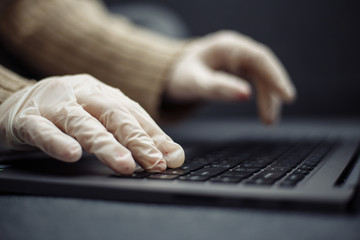 Closeup of young woman working from home during quarantine due to coronavirus pandemia. Beautiful girl stays home wearing medical mask and gloves and typing on a laptop. Covid-19 epidemia concept.