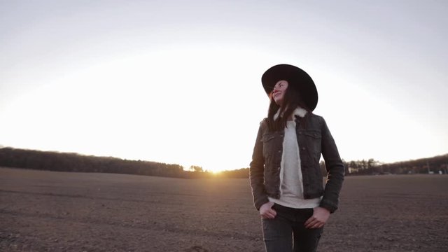 Woman walking lonely through an empty field at sunset wearing cawboy hat and jeans coat. She is smiling and enjoying spring.