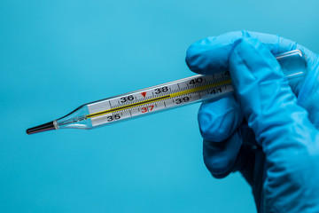 Obraz premium Thermometer in the hand of a woman, gloved hand holding a mercury thermometer for measuring the patients body temperature , view from above , close-up , on a blue background 