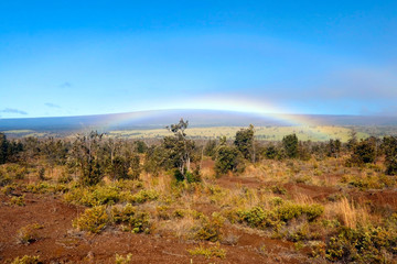 Hawaii Big Island nature background. Scenic landscape with bright blue sky and rainbow over scarce vegetation on lava ground and mountain on the horizon.