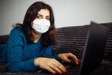 Young businesswoman working from home during quarantine due to coronavirus pandemia. Beautiful girl stays home wearing medical mask and typing on a laptop. Covid-19 epidemia worldwide concept.