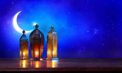 Ramadan lanterns on the table with moon on dark blue background. Beautiful Greeting Card with copy space for Muslim Holidays. An illuminated Arabic lamp. Mixed media.
